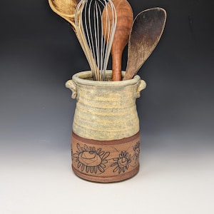 May include: A cylindrical ceramic utensil holder with two small side handles. The top section is a textured cream glaze, while the bottom section is unglazed brown clay featuring a hand-drawn floral pattern. It contains a metal whisk and three wooden spoons.