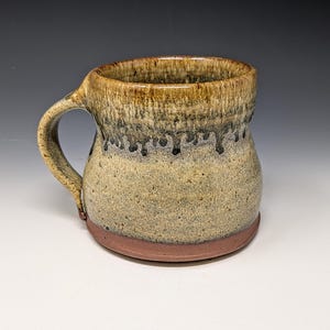 May include: A ceramic mug with a rounded body and a curved handle. The surface features a speckled beige and tan glaze with a dark brown drip pattern cascading from the rim. The base of the mug is a solid, unglazed reddish-brown clay color.
