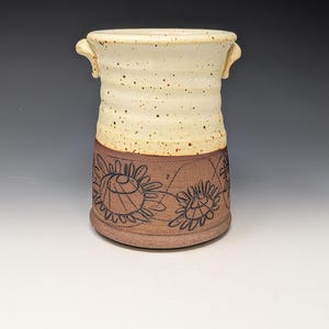 May include: A ceramic utensil holder featuring a cream-colored top section with brown speckles and a brown bottom section decorated with etched black line drawings of flowers. The vessel has two small side handles and a cylindrical shape.