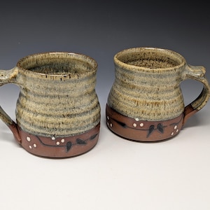 May include: Two ceramic mugs with a speckled beige upper section and a solid reddish-brown base. The lower portion of each mug features a hand-painted dark brown vine pattern with small white dots, and each has a sturdy handle with a thumb rest.