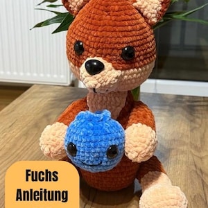 Könnte beinhalten: Ein handgefertigtes Fuchs-Spielzeug aus Häkelarbeit in Orange und Creme, das eine blaue Häkel-Blaubeere hält. Der Fuchs hat schwarze Augen und eine schwarze Nase. Der Text "Fuchs Anleitung PDF" befindet sich auf einem gelben Quadrat.