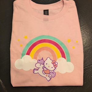 May include: Pink t-shirt with a rainbow, clouds, and a Hello Kitty character riding a unicorn.