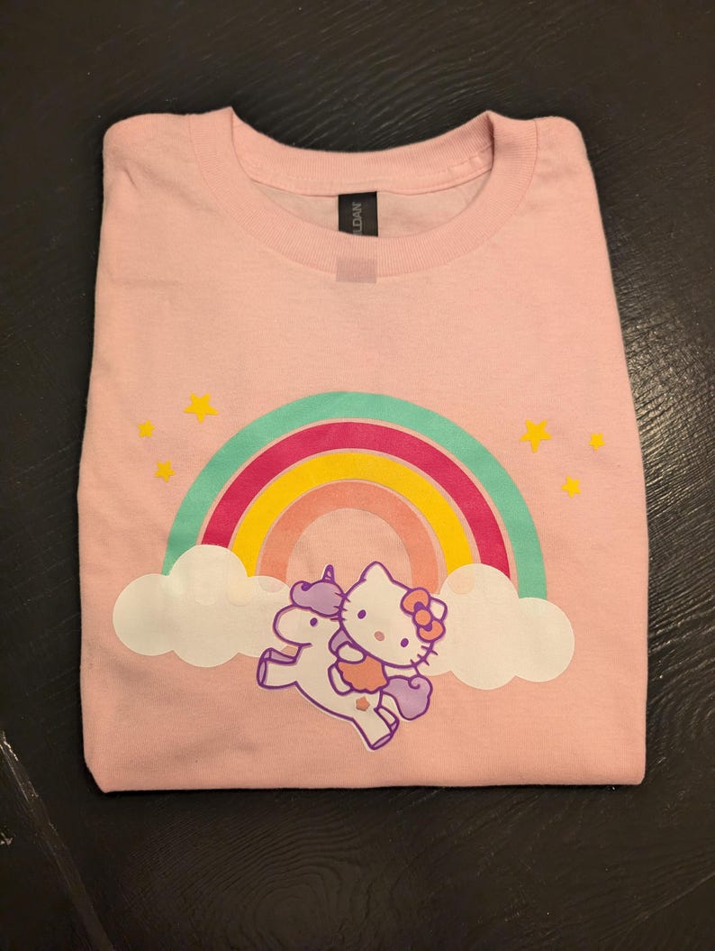 May include: Pink t-shirt with a rainbow, clouds, and a Hello Kitty character riding a unicorn.