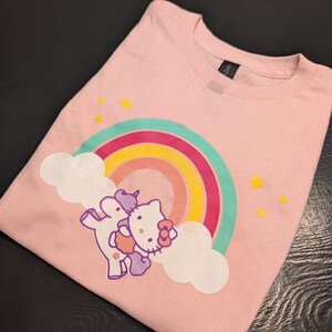 May include: Pink t-shirt with a rainbow and a Hello Kitty unicorn graphic. The unicorn is purple and pink and has a rainbow horn. The rainbow is multi-colored and has clouds on either side.