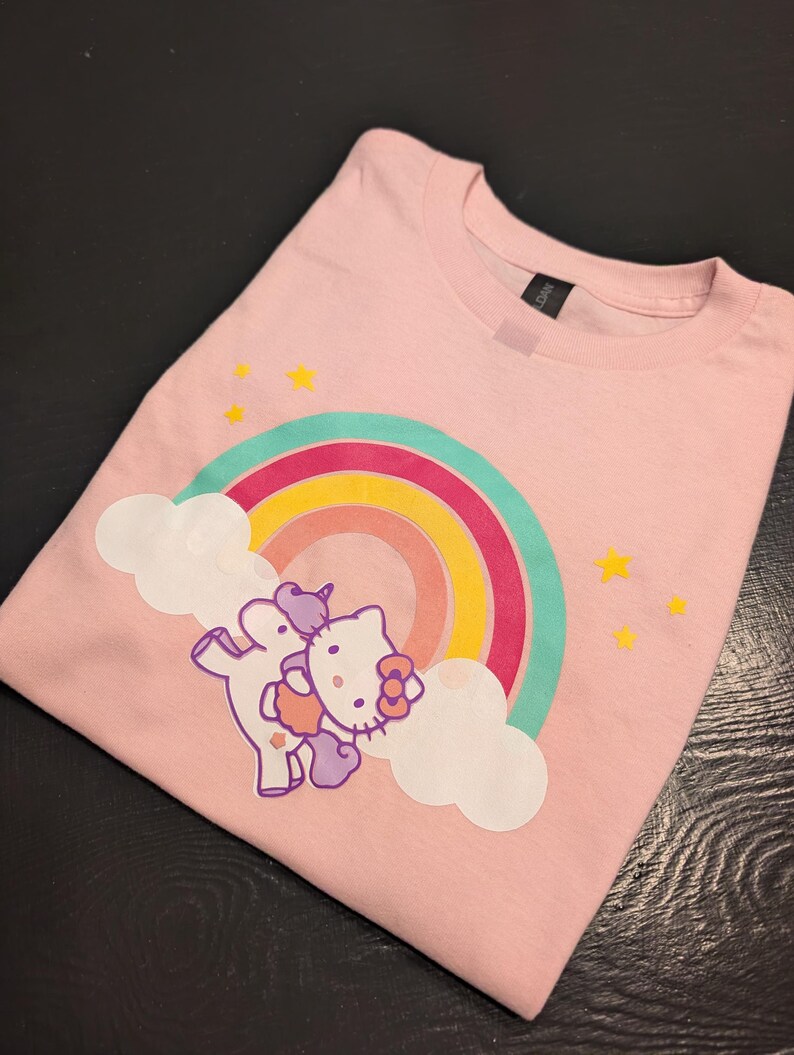 May include: Pink t-shirt with a rainbow and a Hello Kitty unicorn graphic. The unicorn is purple and pink and has a rainbow horn. The rainbow is multi-colored and has clouds on either side.