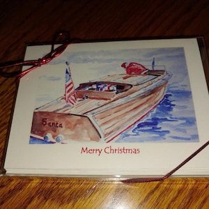 May include: A set of Christmas cards featuring a watercolor illustration of a wooden boat with the word "Santa" on the side and an American flag. The words "Merry Christmas" are printed below the boat. The cards are tied with a red ribbon.