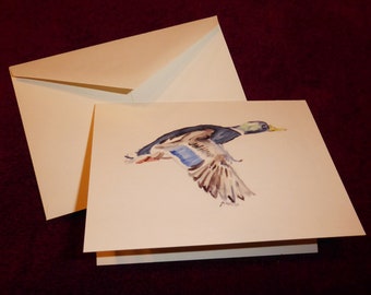 May include: A watercolor painting of a mallard duck in flight. The duck is painted in shades of blue, brown, and white, with a yellow beak. The painting is on a white card with a gold envelope.