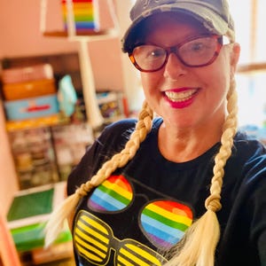 Puede incluir: Una camiseta negra con un estampado gr&aacute;fico de tres pares de gafas de sol, incluyendo un par con lentes de arco&iacute;ris y otro con rayas horizontales amarillas, combinada con una gorra de b&eacute;isbol de camuflaje con un gr&aacute;fico de tigre.