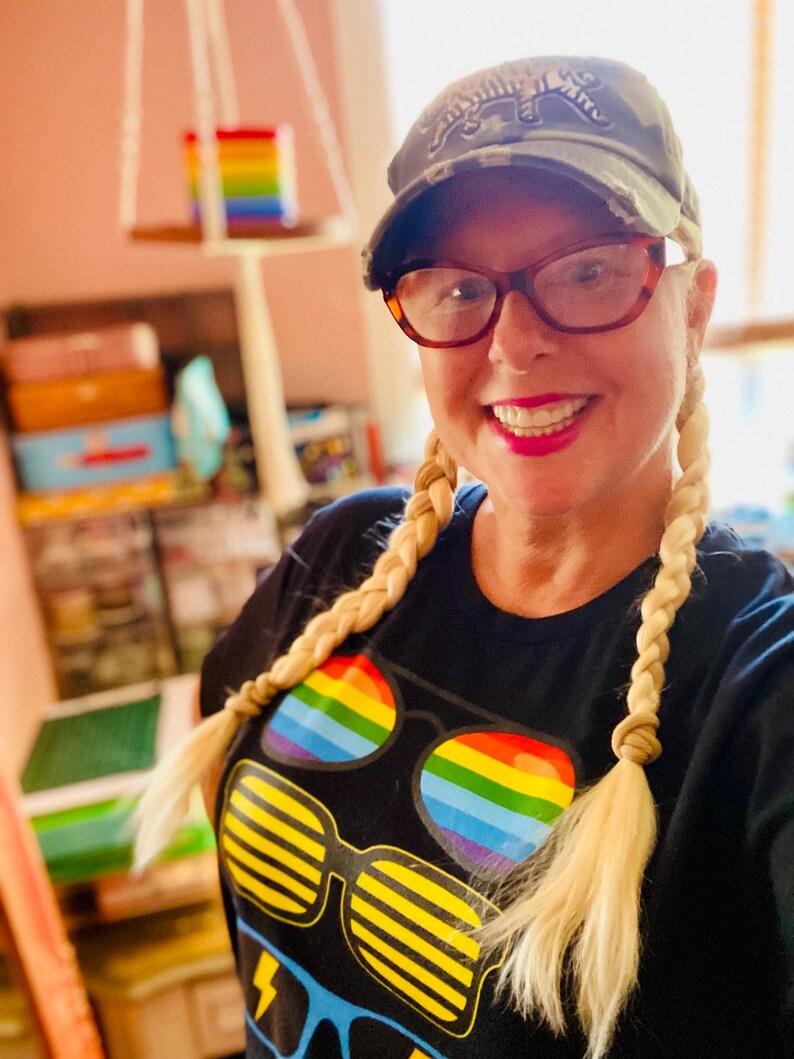 Puede incluir: Una camiseta negra con un estampado gr&aacute;fico de tres pares de gafas de sol, incluyendo un par con lentes de arco&iacute;ris y otro con rayas horizontales amarillas, combinada con una gorra de b&eacute;isbol de camuflaje con un gr&aacute;fico de tigre.