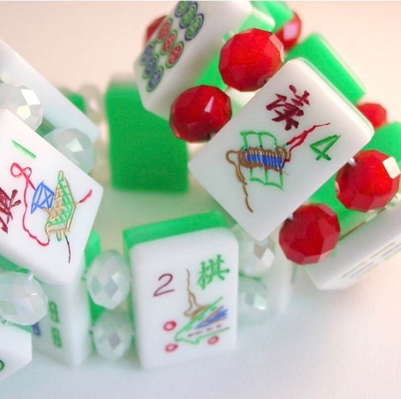 Puede incluir: Una pulsera construida con fichas de mahjong blancas con dise&ntilde;os de caracteres en verde y rojo, intercaladas con cuentas facetadas rojas y transparentes. Las fichas muestran varios s&iacute;mbolos y n&uacute;meros, incluyendo el 2 y el 4.