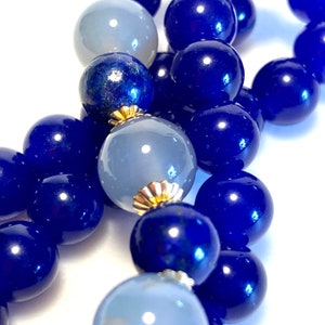 May include: A close-up of a blue and gray beaded bracelet. The bracelet is made of round beads, some of which are blue and some of which are gray. The beads are strung together on a clear string.