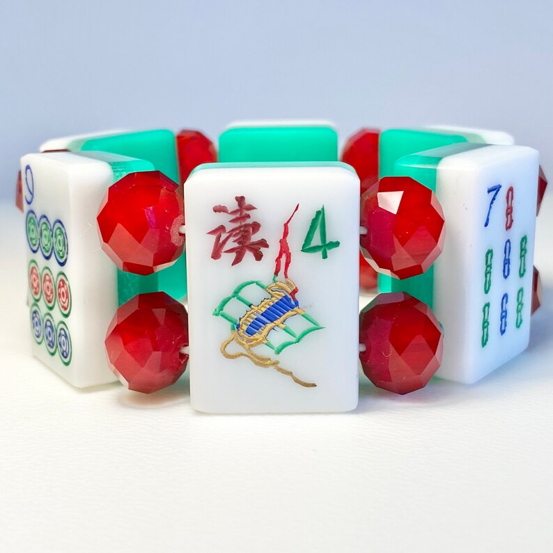 Puede incluir: Una pulsera construida con fichas de Mahjong blancas que presentan varios s&iacute;mbolos y n&uacute;meros grabados en colores, intercaladas con cuentas rojas facetadas y transl&uacute;cidas, unidas por espaciadores rectangulares de color verde claro.