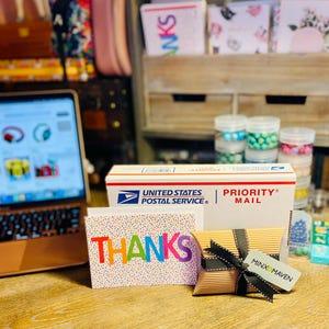 Puede incluir: Una caja de env&iacute;o blanca de United States Postal Service Priority Mail, una tarjeta de felicitaci&oacute;n blanca con puntos de confeti coloridos y la palabra THANKS en letras arco&iacute;ris, y una peque&ntilde;a caja almohada marr&oacute;n con una cinta negra y una etiqueta Minx & Maven.