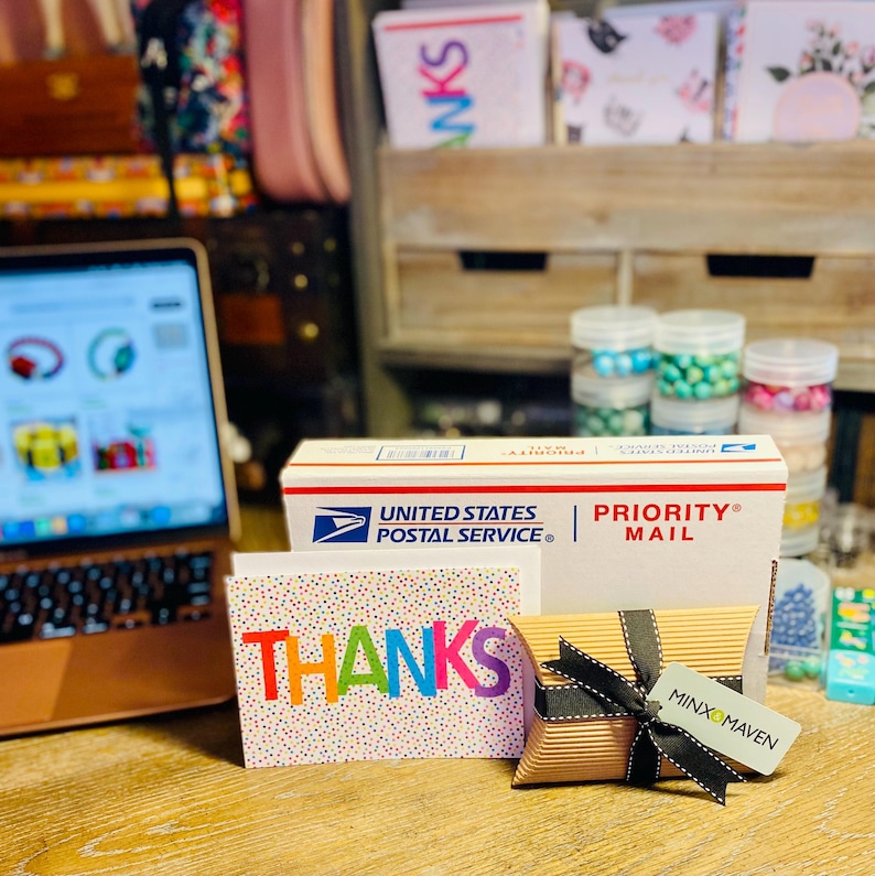 Puede incluir: Una caja de env&iacute;o blanca de United States Postal Service Priority Mail, una tarjeta de felicitaci&oacute;n blanca con puntos de confeti coloridos y la palabra THANKS en letras arco&iacute;ris, y una peque&ntilde;a caja almohada marr&oacute;n con una cinta negra y una etiqueta Minx & Maven.