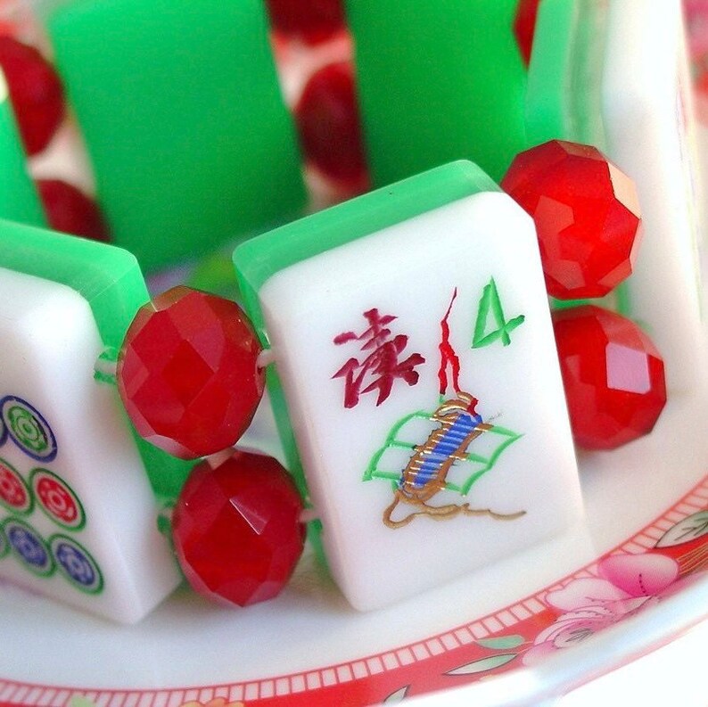 Puede incluir: Una pulsera hecha de fichas de mahjong blancas y verdes intercaladas con cuentas rojas facetadas y transl&uacute;cidas. Las fichas presentan caracteres y s&iacute;mbolos grabados en rojo, verde y azul, incluyendo el n&uacute;mero 4 y caligraf&iacute;a china.