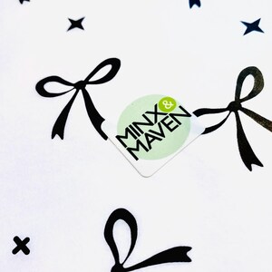 Puede incluir: Una tela blanca con un patr&oacute;n repetitivo de lazos negros y peque&ntilde;as estrellas de cuatro puntas. En el centro hay una pegatina cuadrada de esquinas redondeadas con el texto MINX & MAVEN en negro sobre un fondo circular de color verde claro.