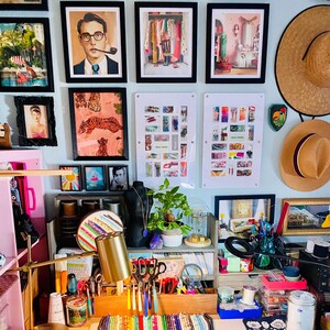 Puede incluir: Una pared con m&uacute;ltiples l&aacute;minas enmarcadas, dos paneles acr&iacute;licos blancos con objetos coloridos y dos sombreros de paja. Debajo, un organizador de escritorio de madera contiene herramientas, cuentas, una planta y una l&aacute;mpara.