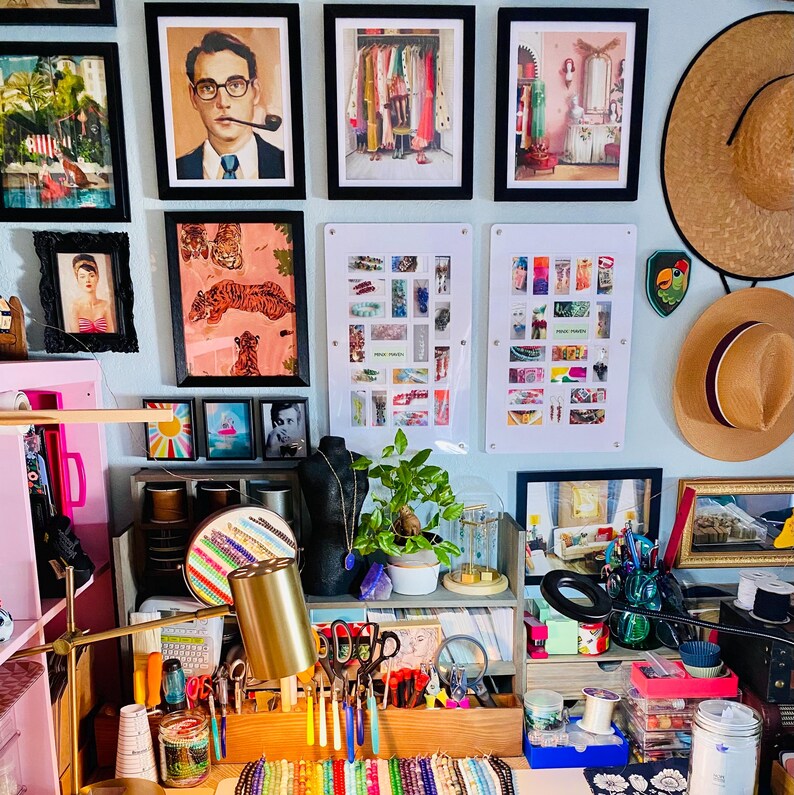 Puede incluir: Una pared con m&uacute;ltiples l&aacute;minas enmarcadas, dos paneles acr&iacute;licos blancos con objetos coloridos y dos sombreros de paja. Debajo, un organizador de escritorio de madera contiene herramientas, cuentas, una planta y una l&aacute;mpara.