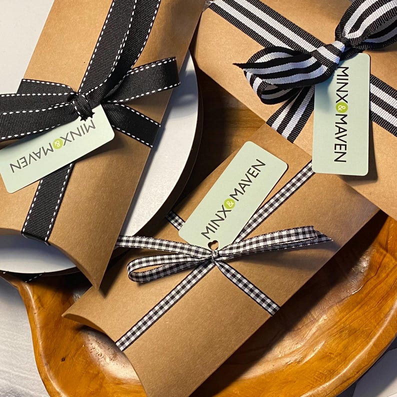 Puede incluir: Tres cajas de cart&oacute;n marr&oacute;n con forma de almohada atadas con cintas de rayas o cuadros en blanco y negro. Cada caja tiene una etiqueta rectangular de color verde claro con el texto MINX & MAVEN impreso en negro y un peque&ntilde;o c&iacute;rculo verde.