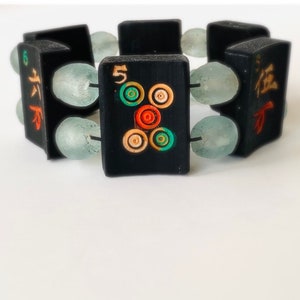 May include: A black bracelet featuring rectangular wooden beads with colorful circular designs and Chinese characters. The beads are interspersed with light blue, oval-shaped beads. The bracelet is displayed on a white surface.