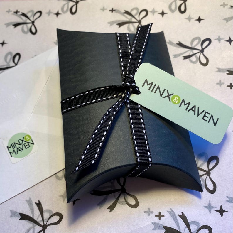 Puede incluir: Una caja tipo almohada negra atada con una cinta negra con costuras blancas. Una etiqueta rectangular de color verde claro unida a la cinta muestra el texto MINX & MAVEN en negro. Se incluye un sobre blanco a juego con una peque&ntilde;a pegatina verde MINX & MAVEN.