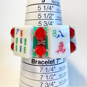 Puede incluir: Una pulsera con fichas de Mahjong rectangulares blancas con caracteres grabados en verde, azul y rojo, alternadas con cuentas rojas facetadas. La pulsera se muestra en un mandril c&oacute;nico blanco con una circunferencia de 17,8 cm.