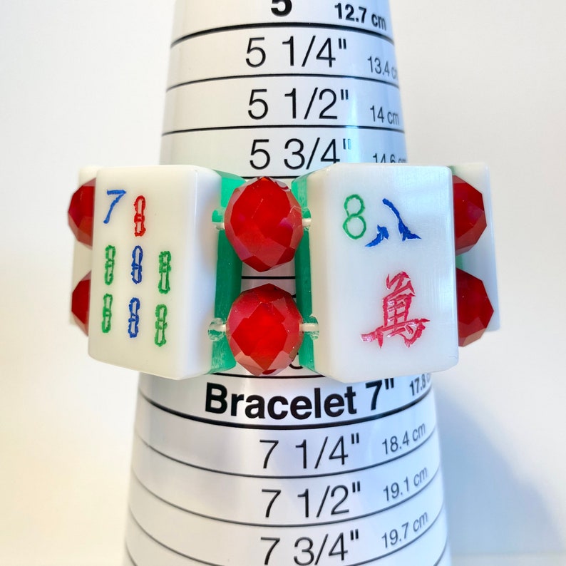 Puede incluir: Una pulsera con fichas de Mahjong rectangulares blancas con caracteres grabados en verde, azul y rojo, alternadas con cuentas rojas facetadas. La pulsera se muestra en un mandril c&oacute;nico blanco con una circunferencia de 17,8 cm.