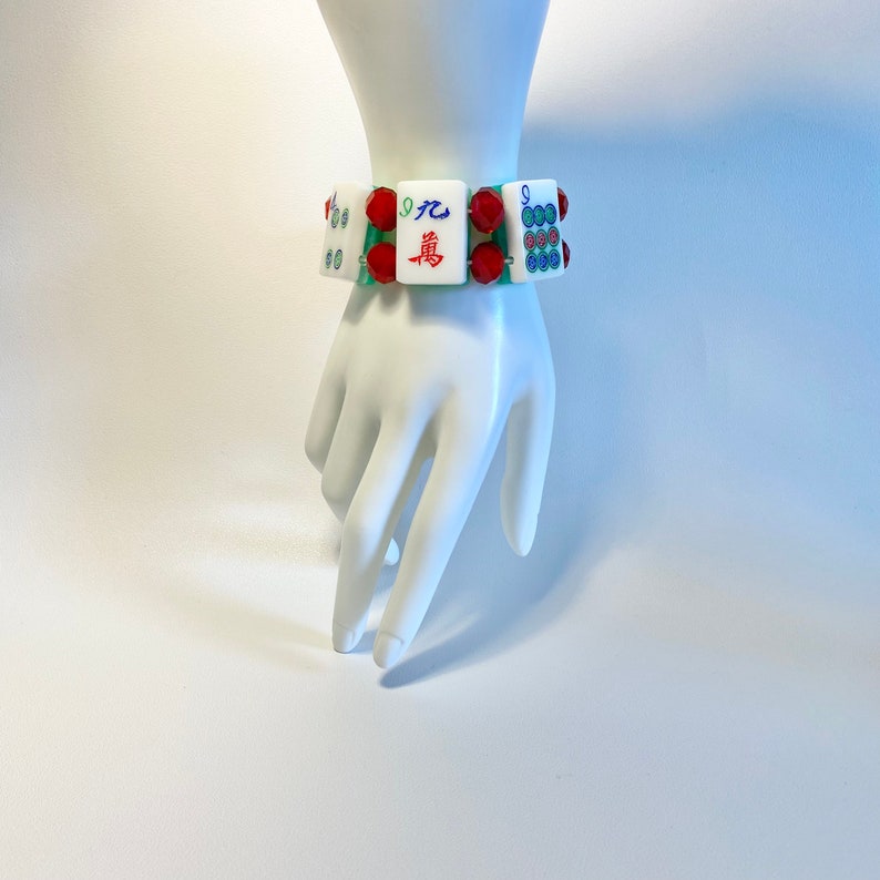 Puede incluir: Una pulsera compuesta por fichas de mahjong rectangulares blancas con caracteres chinos y s&iacute;mbolos circulares de colores, intercaladas con cuentas rojas facetadas y unidas por un cord&oacute;n el&aacute;stico verde, formando un accesorio para la mu&ntilde;eca.