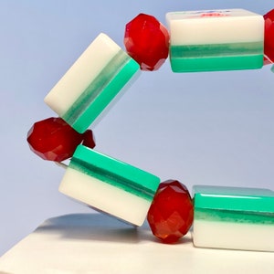 Puede incluir: Una pulsera con piezas rectangulares blancas y verdes que alternan con cuentas rojas transl&uacute;cidas facetadas. Las piezas se asemejan a fichas de mahjong, con una pieza visible que muestra un car&aacute;cter rojo y verde. Cuentas redondas y pulidas.