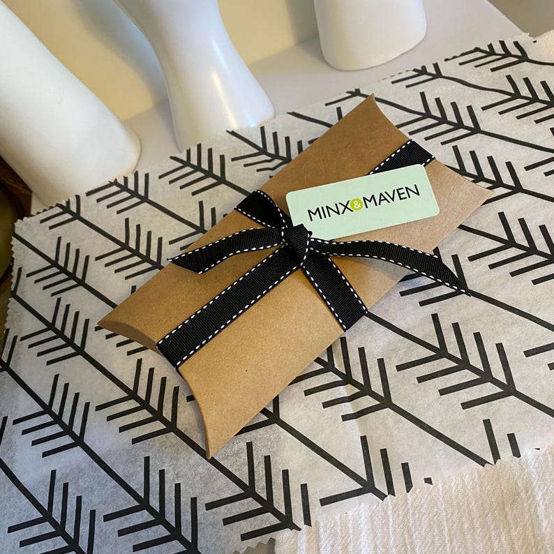 Puede incluir: Una caja tipo almohada de papel kraft marr&oacute;n atada con una cinta de grosgrain negra con bordes cosidos en blanco. Una etiqueta rectangular verde claro est&aacute; metida bajo la cinta, mostrando el texto negro y verde MINX & MAVEN en letra sans-serif.