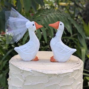 May include: Two white goose figurines with orange beaks and feet positioned on top of a frosted cake. The left goose wears a white tulle veil with a small flower, and the right goose wears a small black bow tie around its neck.