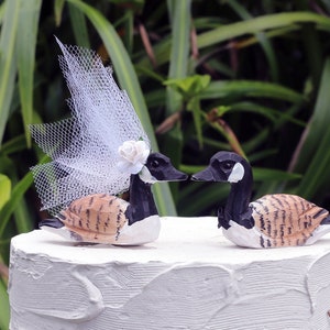 May include: A white wedding cake with a pair of wooden goose cake toppers. The female goose is wearing a white tulle veil and a white flower. The male goose is wearing a black bow tie.