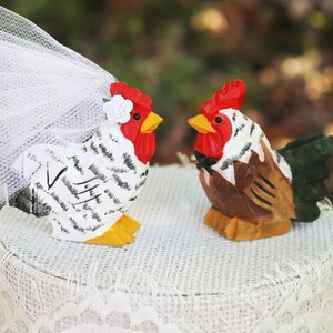 May include: Wooden rooster and hen cake toppers. The hen is white with black markings and a veil. The rooster is brown and tan with a black bow tie. Both have red combs and yellow beaks. A unique wedding cake decoration.