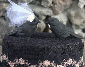 May include: Black wooden bird wedding cake topper with a white tulle veil and a white flower on the bride's head. The birds are perched on a black lace cake stand.