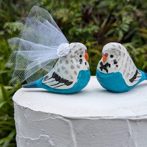 May include: Two hand-painted wooden parakeet figurines, one adorned with a white veil and flower, sit atop a white cake. The birds feature blue, white, and black details, with orange beaks. The figurines are a charming wedding cake topper.