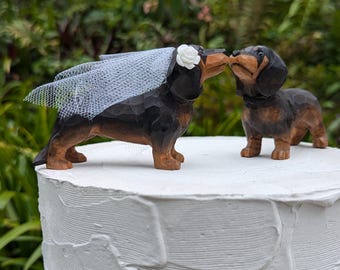 May include: A wooden dachshund wedding cake topper set. The bride dachshund is wearing a white veil and a white flower. The groom dachshund is looking at the bride and they are kissing.