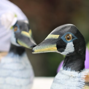 May include: Two wooden painted geese figurines. The goose on the right is wearing a white veil and has a yellow beak and black and white painted feathers. The goose on the left is out of focus.