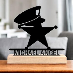 Può includere: Silhouette nera di un poliziotto a forma di stella, montata su una base di legno. Il nome "MICHAEL ANGEL" è visualizzato in testo nero sotto la stella. La statuetta è esposta su un tavolo di legno.