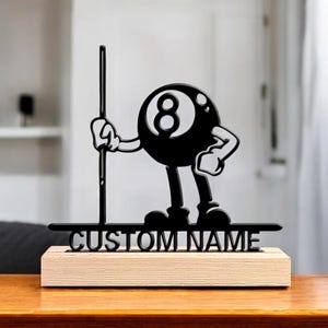 May include: Black metal figure of a billiard ball with the number 8, holding a pool cue. It's mounted on a light wood base with the text "CUSTOM NAME". The background is blurred, showing a wooden table and a white curtain.