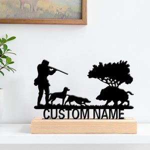 Peut inclure: Une silhouette en métal noir représentant un chasseur avec un fusil, deux chiens et un sanglier sous un arbre, fixée sur un socle en bois clair. Le socle porte l'inscription CUSTOM NAME en lettres capitales noires sur le devant.