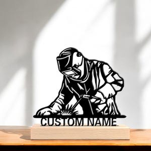 May include: Black metal sculpture of a welder in action, wearing a welding helmet and holding a torch. The sculpture is mounted on a light-colored wooden base with the text "CUSTOM NAME".