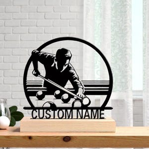 May include: Black metal silhouette of a billiards player with a cue, balls, and the text "CUSTOM NAME" on a wooden base. The design is set within a circular frame, suitable for a personalized gift.
