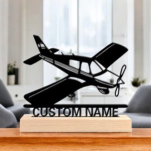 Op de afbeelding: Zwart metalen vliegtuigsilhouet op een houten voet. Het vliegtuig heeft gedetailleerde vleugels, een propeller en een staart. De basis heeft de tekst "CUSTOM NAME" in het zwart. Het item wordt op een houten tafel weergegeven.