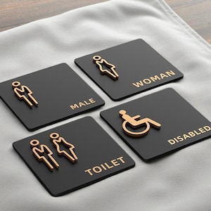 Puede incluir: Cuatro letreros de baño cuadrados negros con símbolos y texto dorados. Los letreros indican acceso para hombres, mujeres, inodoros y discapacitados. Los letreros están dispuestos sobre una superficie gris.