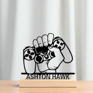 May include: A black and white gaming controller design, held in a fist, mounted on a wooden base. The text "ASHTON HAWK" is displayed below the controller. The background is a white curtain.