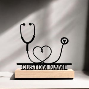 Pode incluir: Escultura de estetoscópio em metal preto com design em forma de coração, montada numa base de madeira clara. O texto "CUSTOM NAME" está na base. Um item decorativo.
