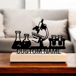 Può includere: Decorazione da scrivania nera tagliata al laser con microscopio, becher e provette. La base è in legno chiaro, con le parole "CUSTOM NAME" ritagliate nel design nero. Un regalo unico per gli appassionati di scienza.