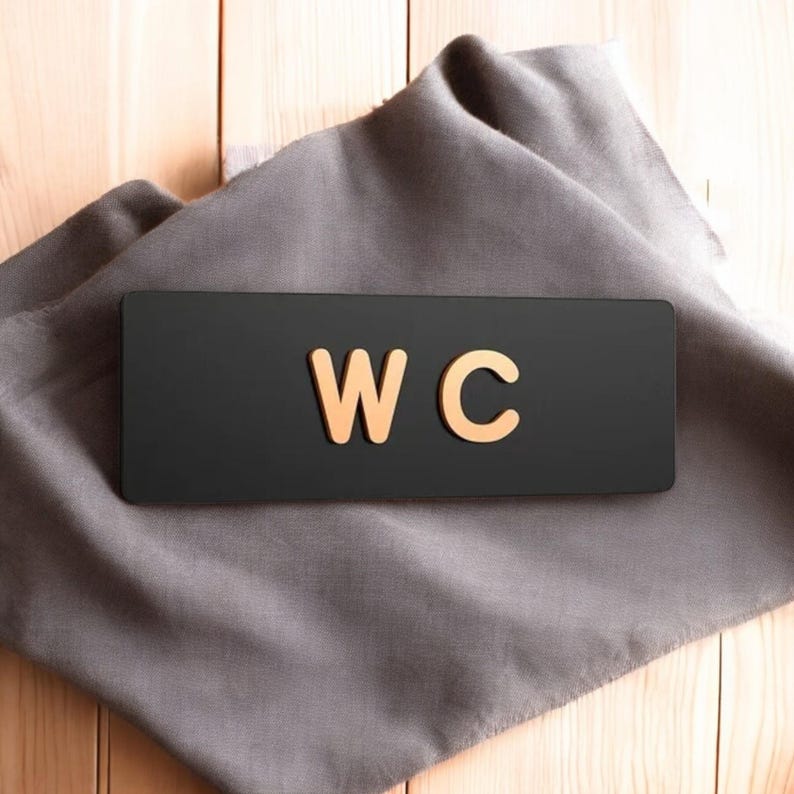 May include: A black rectangular sign with the gold letters "WC" on a gray cloth. The sign is likely for a restroom or washroom. The background is a light-colored wooden surface.