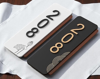 Könnte beinhalten: Zwei rechteckige Türschilder mit der Nummer "2008". Ein Schild ist weiß mit schwarzen Zahlen und einem Lotusblumen-Design, das andere ist schwarz mit goldenen Zahlen und einem goldenen Wellen-Design. Beide haben einen Holzsockel.