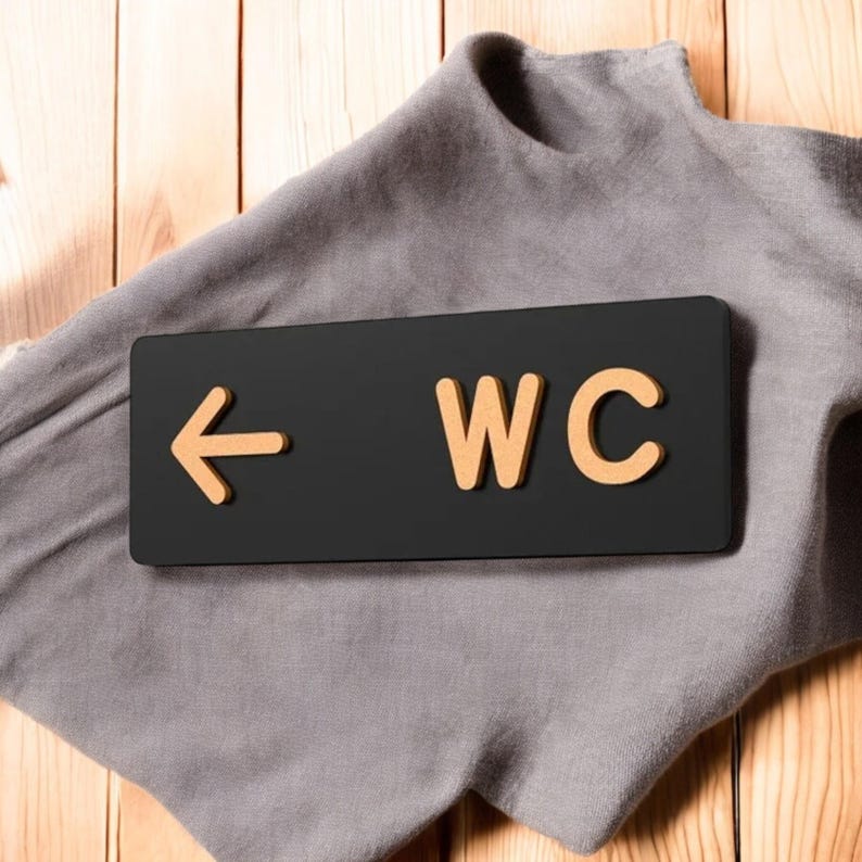 May include: A black rectangular sign with the text "WC" in a light brown color and an arrow pointing left. The sign is on a gray cloth, with a wooden background.